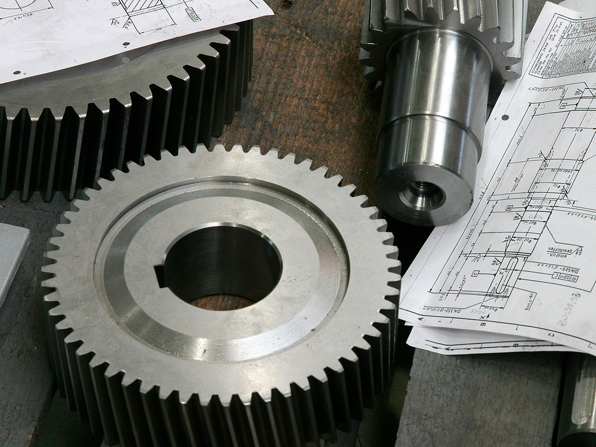 Einzelteile mit technischen Bauzeichnungen von KACHELMANN GETRIEBE GmbH Die Einzelteile der Firma KACHELMANN GETRIEBE GmbH liegen mit einigen technischen Zeichnungen auf einer Werkbank, die eine dunkelbraune Holzplatte besitzt. Die Einzelteile des Getriebes sind auseinander gebaut und liegen auf der Werkbank. KACHELMANN GETRIEBE GmbH